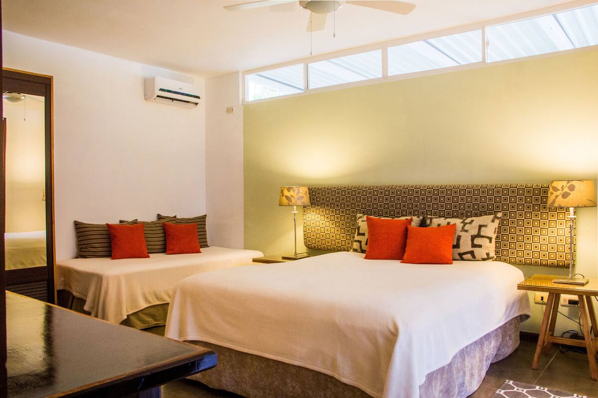 A hotel room with two beds, a ceiling fan, an air conditioning unit, and a view through the window.