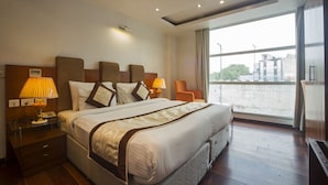 A hotel room with a large bed, two bedside tables with lamps, a view of the city through a large window, and a modern ceiling design. - Trimrooms Palm D'or (New Delhi)