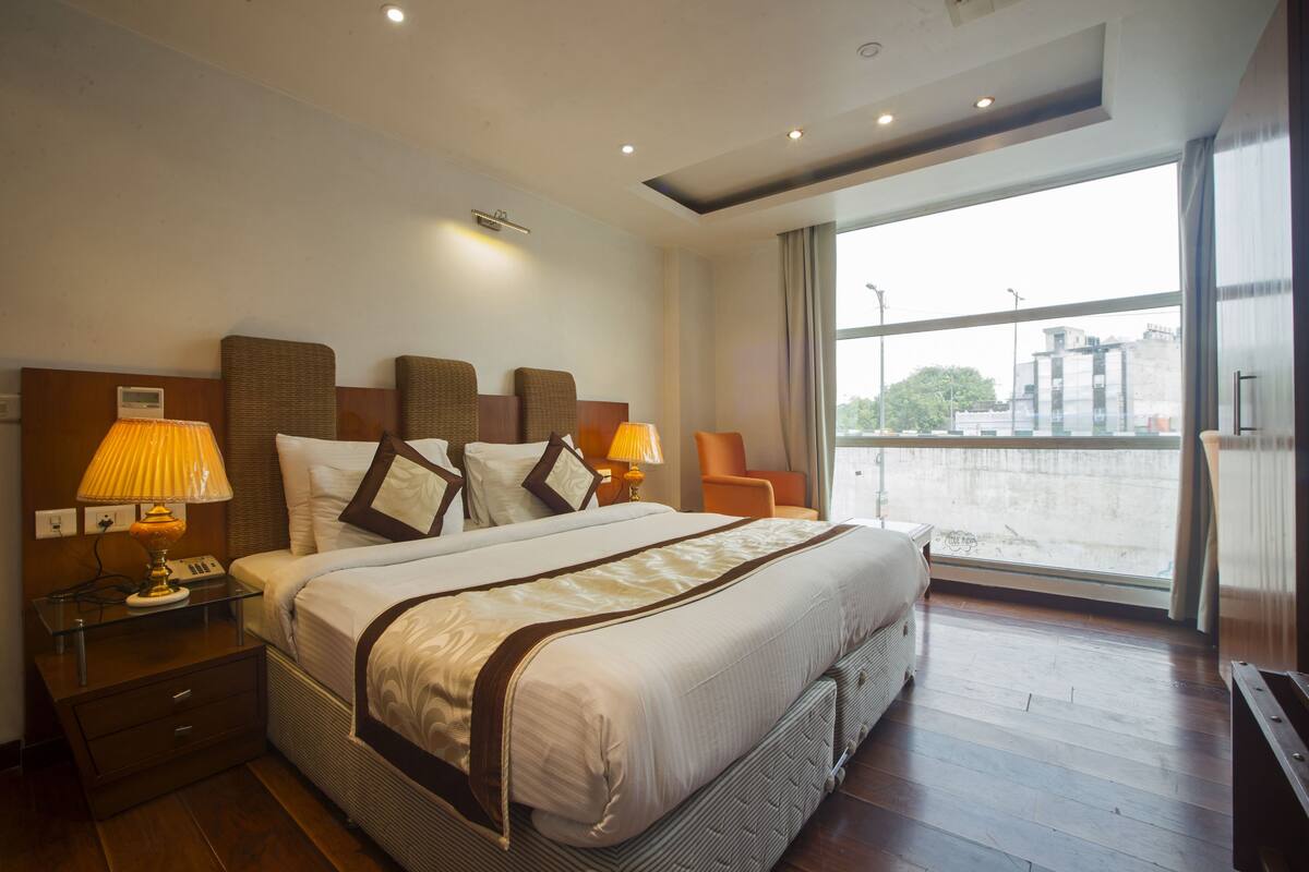 Habitación de hotel con una cama grande, dos mesitas de noche con lámparas, vista a la ciudad a través de un ventanal amplio y un diseño de techo moderno.