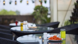 A glass table with a plate of food, a salt shaker, and a glass of yellow liquid, likely juice, in an outdoor setting with wicker furniture and string lights in the background. - Trimrooms Palm D'or (New Delhi)