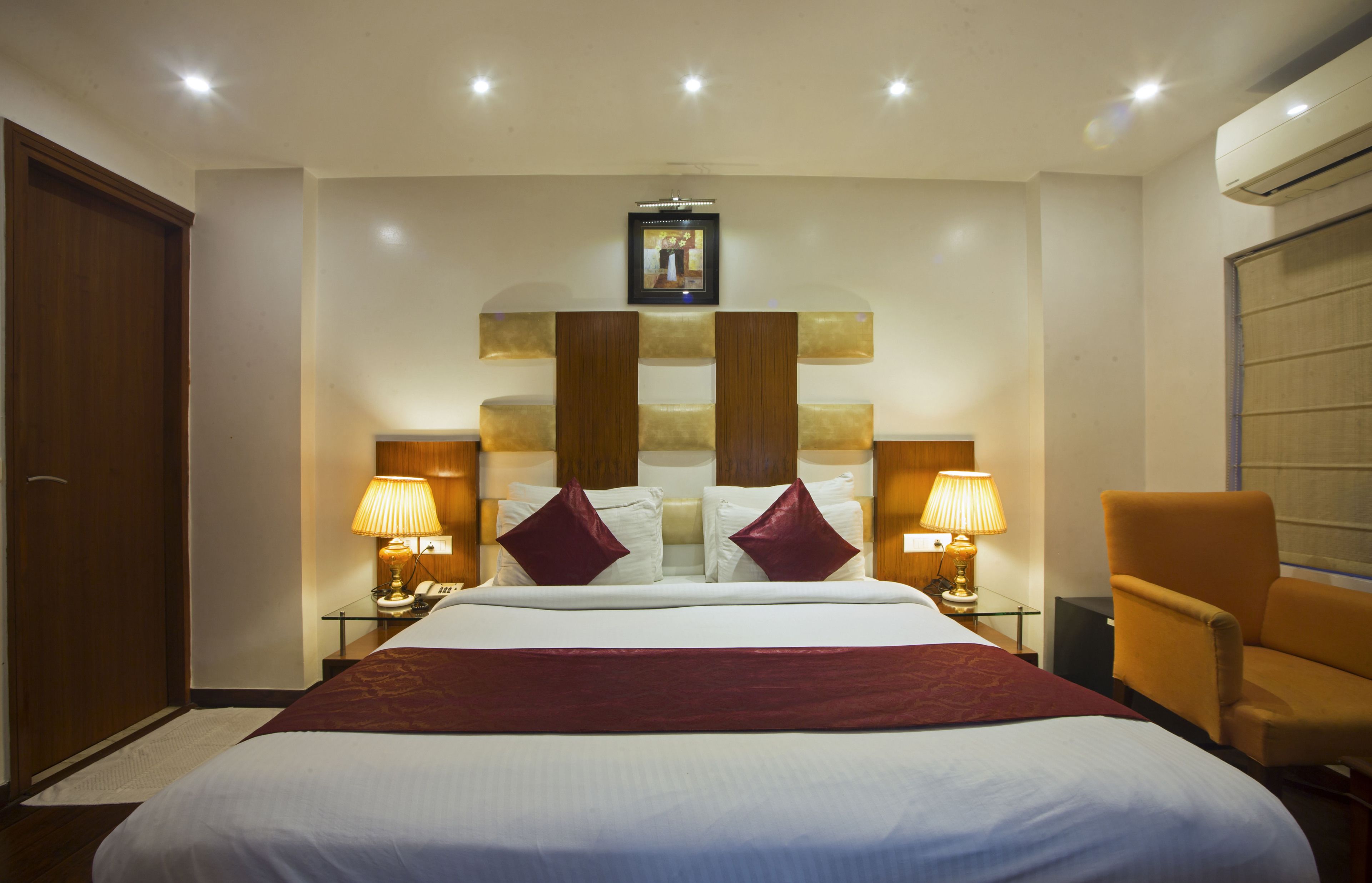 A hotel room with a large bed, two bedside lamps, a wooden headboard, a picture above the bed, and an air conditioning unit.