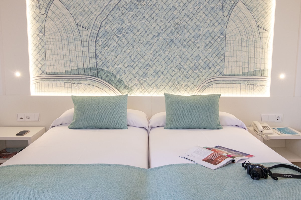 Une chambre d’hôtel avec un grand lit, deux oreillers, une table de chevet avec un téléphone et un magazine, et un luminaire fixé au mur.
