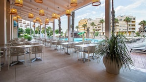 Un amplio espacio exterior con mesas y sillas, lámparas de mimbre colgantes, y vista a una piscina y palmeras.