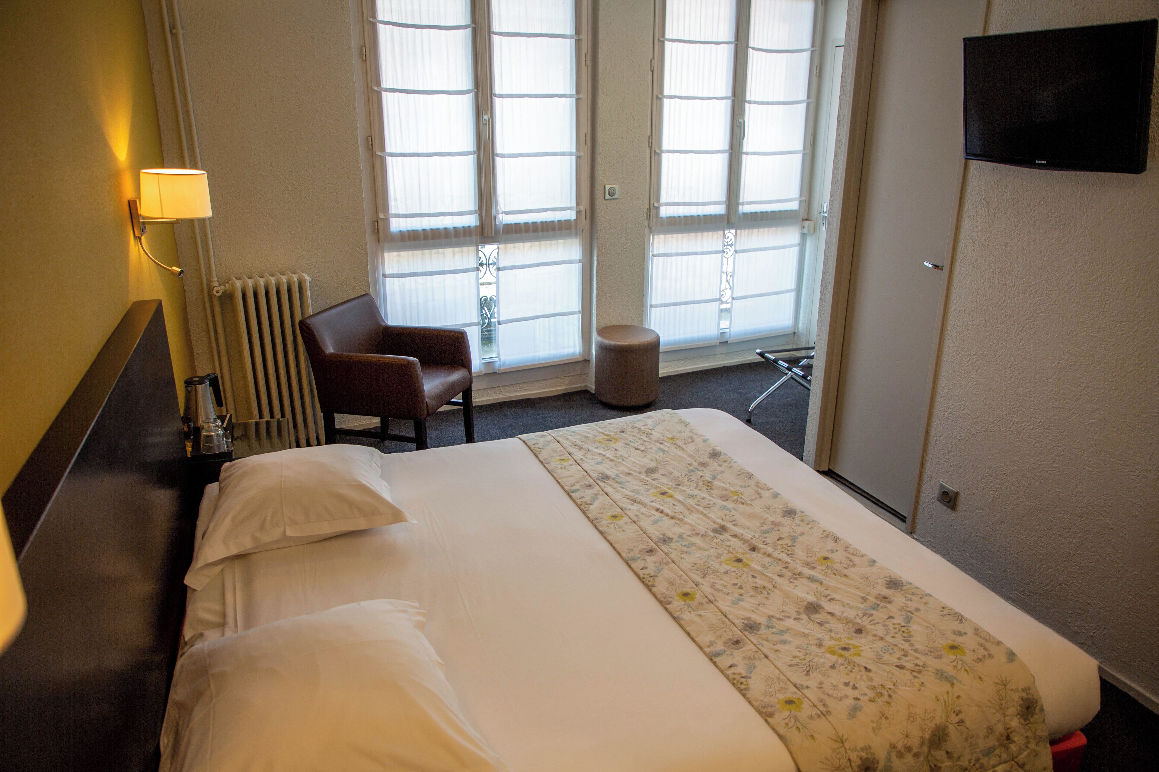 A hotel room with a bed, a chair, a small table, a lamp, a radiator, a television, and large windows with blinds.