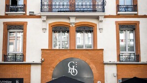 Een gebouw met een grote bogenpoort, terras met parasols en een zichtbare gevel met een bord.