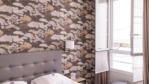Een slaapkamer met bloemenbehang, een grijs gestoffeerd hoofdeinde, een wit bed met twee kussens, een nachtkastje met een lamp en een raam met gordijnen.