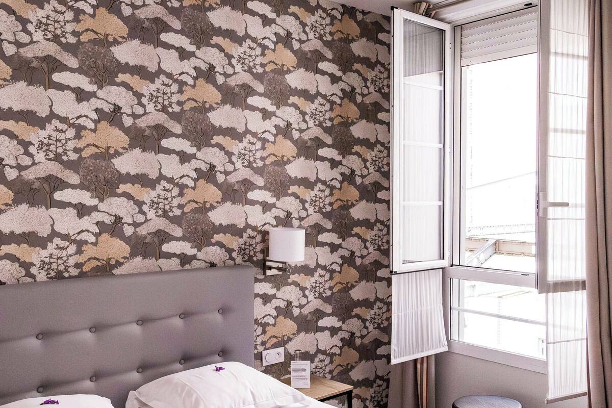 Een slaapkamer met bloemenbehang, een grijs gestoffeerd hoofdeinde, een wit bed met twee kussens, een nachtkastje met een lamp en een raam met gordijnen.