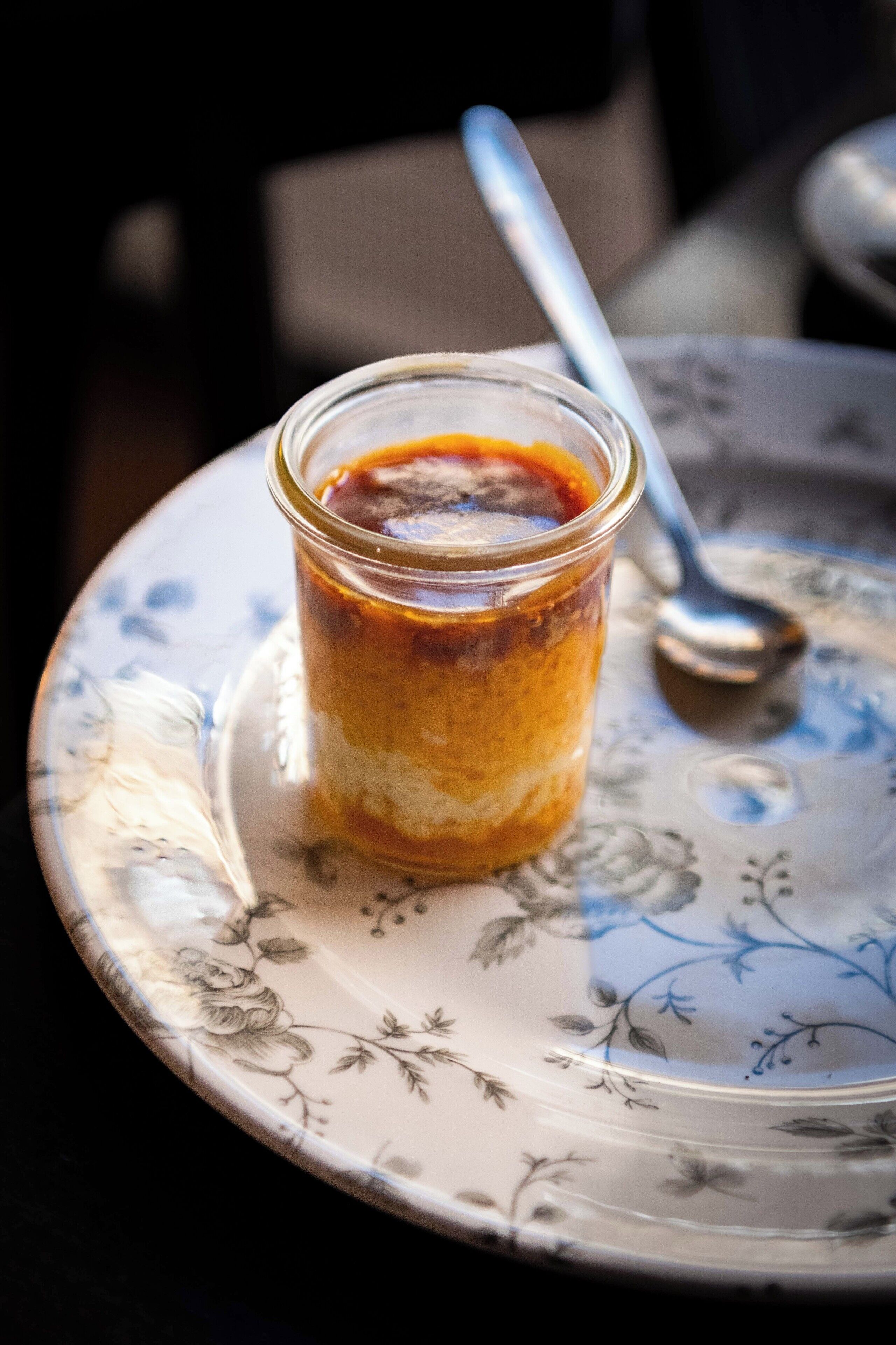 A dessert in a glass container on a floral plate with a spoon.