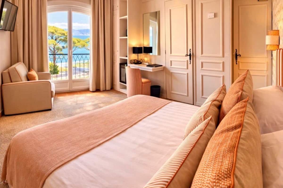 A hotel room with a large bed, a desk, a chair, a TV, and a balcony with a view.