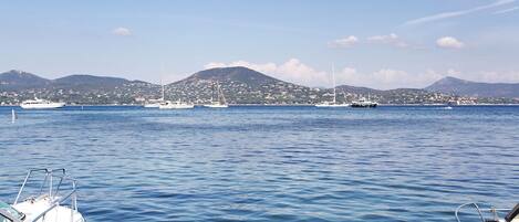 A marina with boats docked, calm water, and a distant view of a coastal town with mountains.