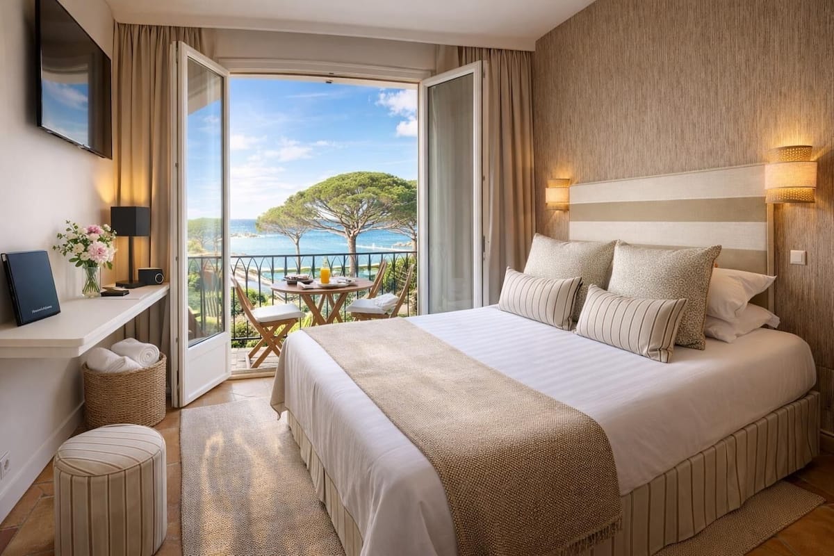 A hotel room with a large bed, a desk, a chair, a small table with a view, and a balcony with a table and chairs.
