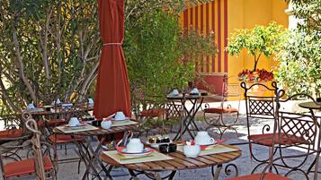 Un café al aire libre con mesas y sillas, rodeado de vegetación y un edificio amarillo.