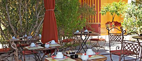 An outdoor café with tables and chairs, surrounded by greenery and a yellow building.