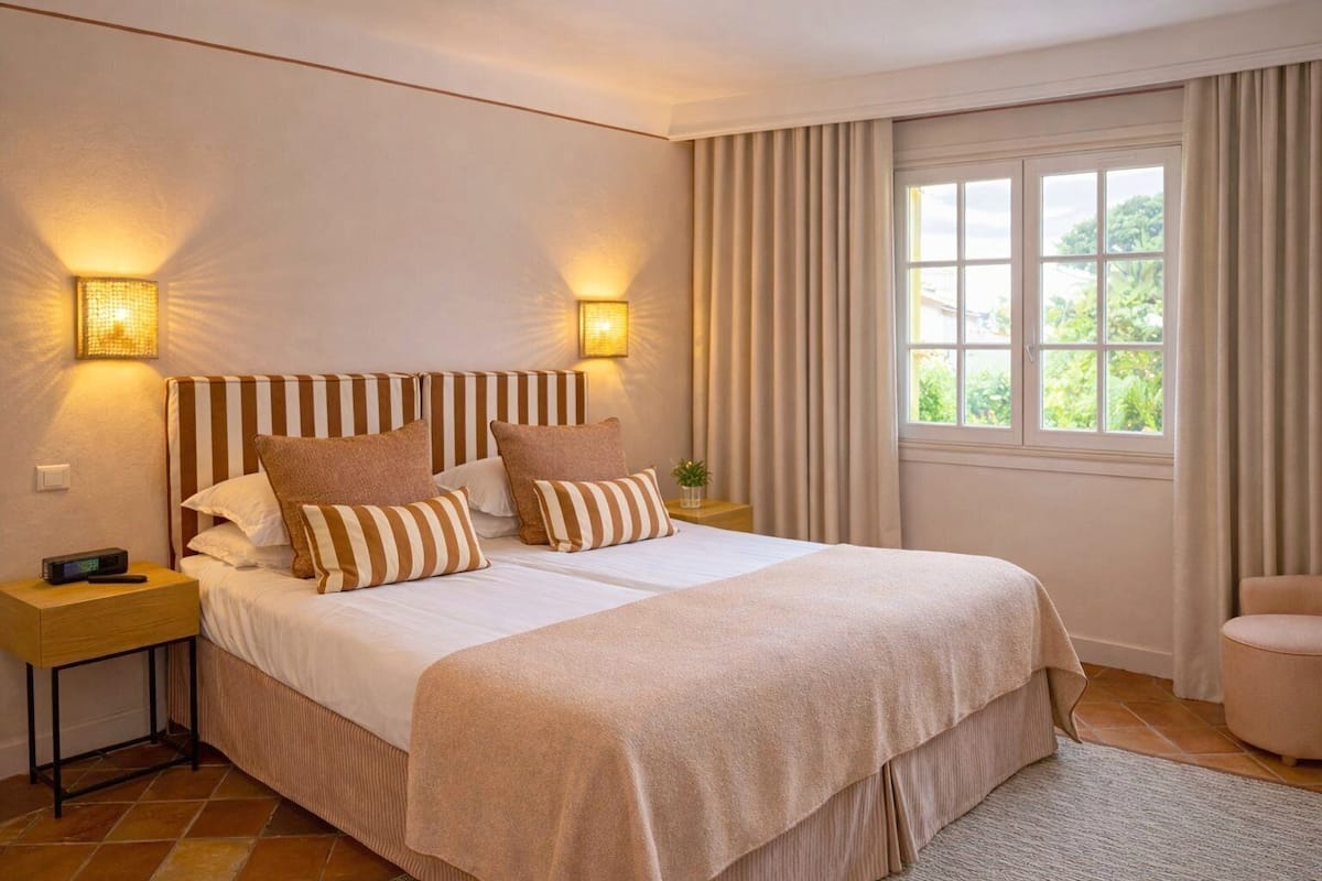 A double bed with striped headboard, two bedside lamps, a nightstand, a window with curtains, and a chair.