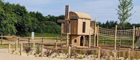 A wooden playhouse with a rope fence, surrounded by greenery and a playground area.