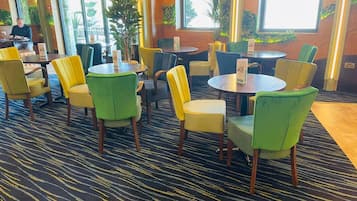 Un hall de hotel con mesas y sillas redondas, ventanas amplias y plantas de interior.