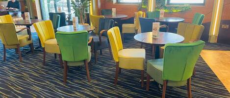 A hotel lobby with round tables and chairs, large windows, and indoor plants.