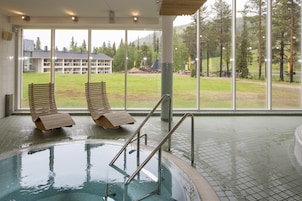 Área de piscina com dois espreguiçadeiras de madeira e vista para um edifício e árvores através de grandes janelas.