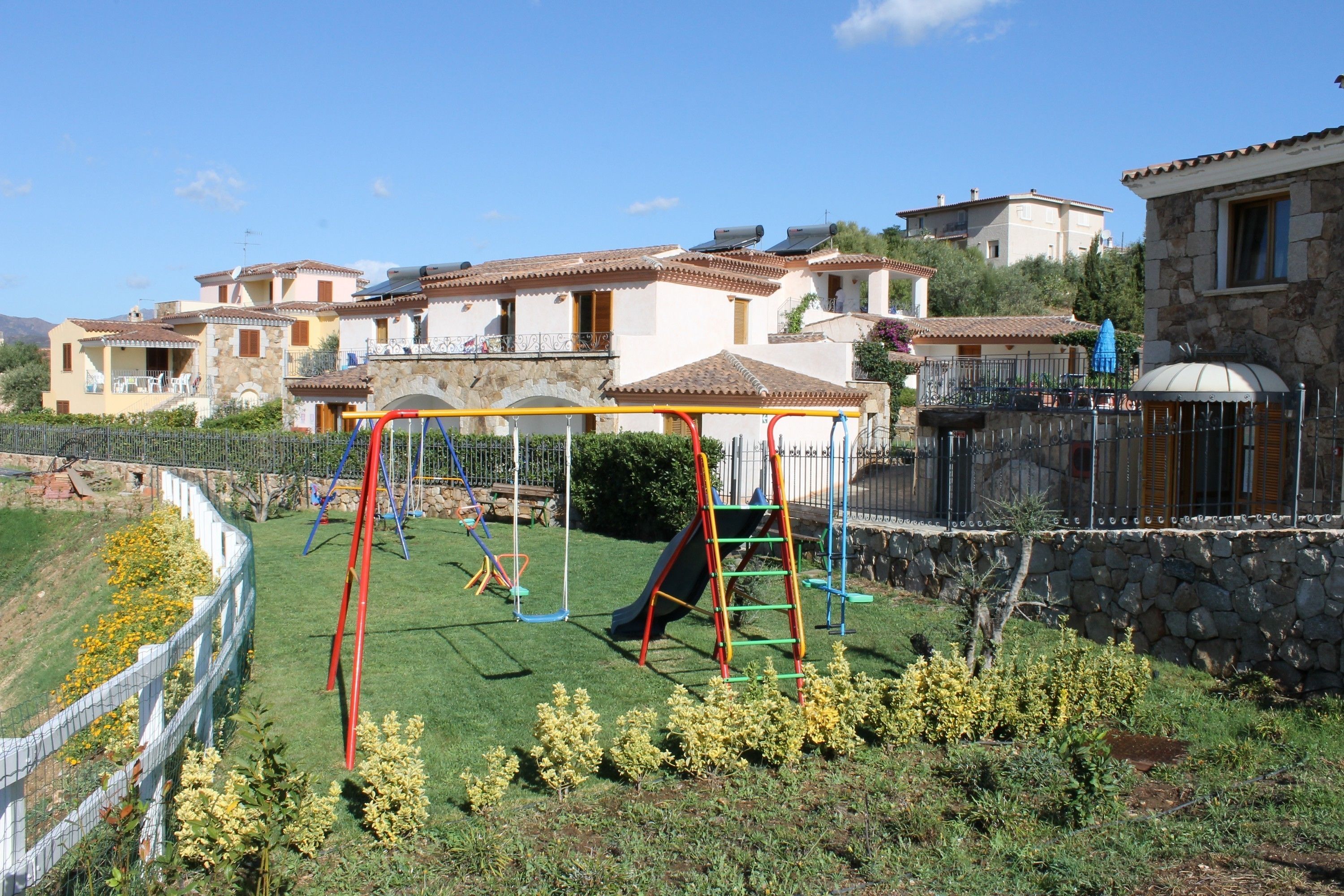 A playground with a slide and swings, surrounded by a stone wall and a fence.