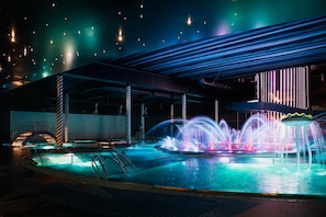 A nighttime scene of a modern building with a pool area featuring illuminated water jets and a unique architectural design with a sloped roof and vertical light strips.