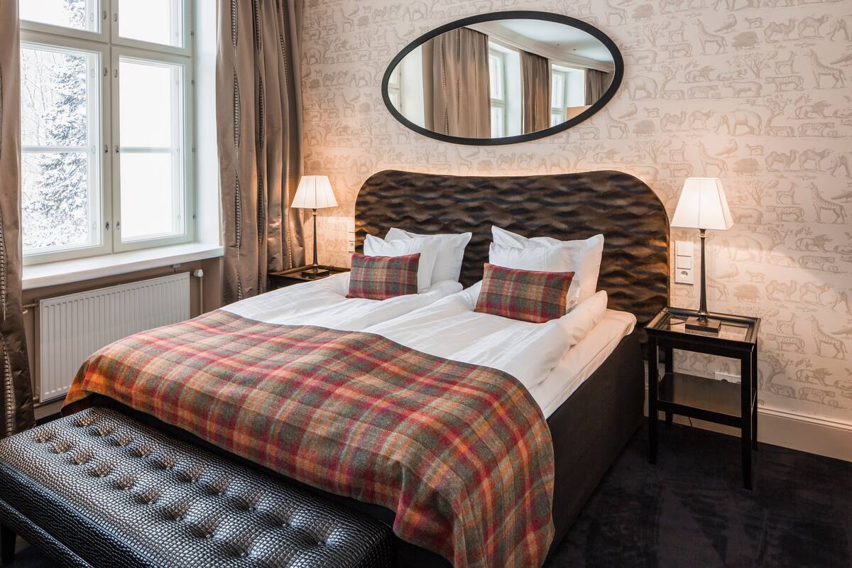 Une chambre à coucher avec un grand lit, deux tables de chevet avec des lampes, un grand miroir rond au-dessus du lit et une fenêtre avec des rideaux.