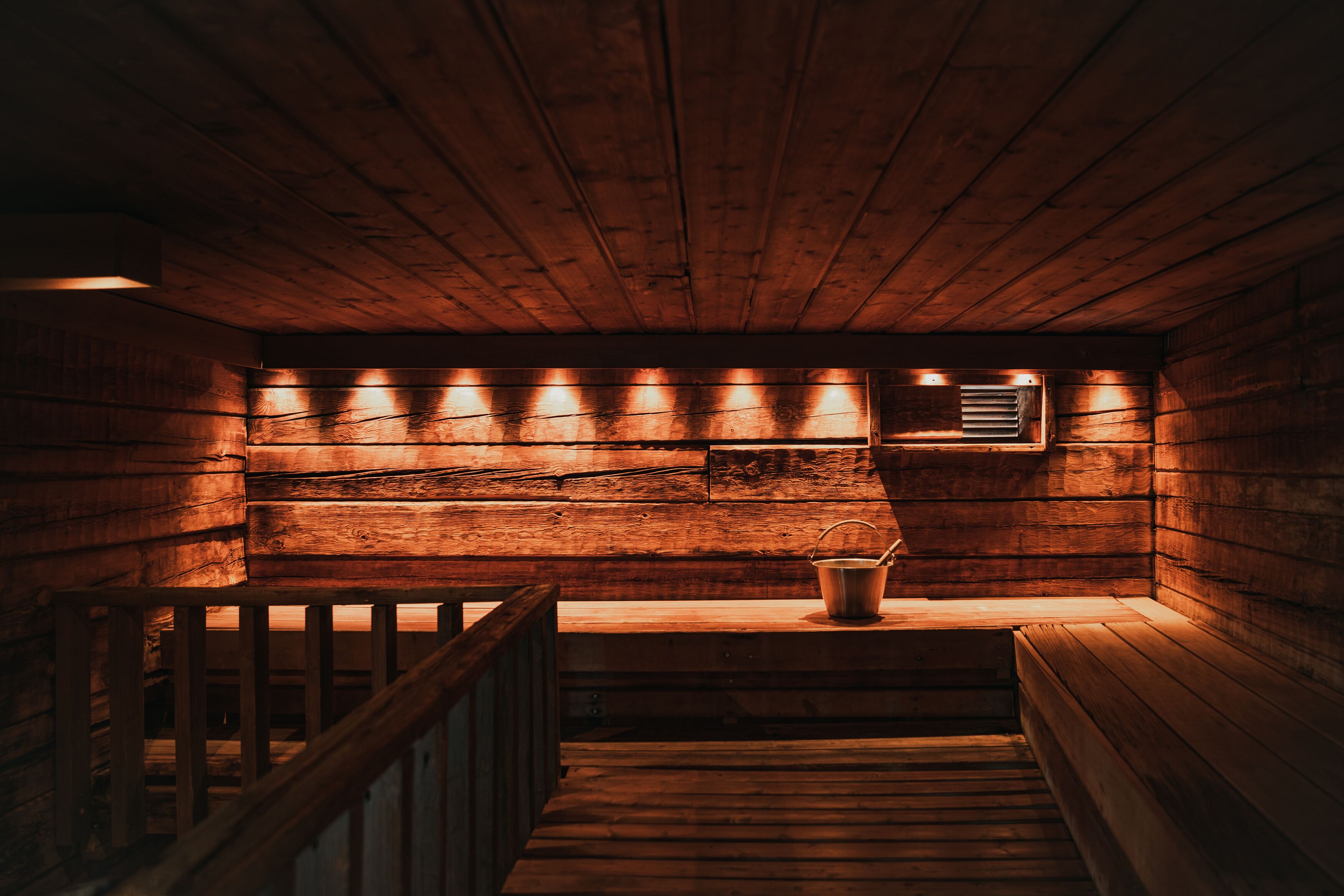Interior de una cabaña de madera con escalera de madera, un banco y un balde de metal.