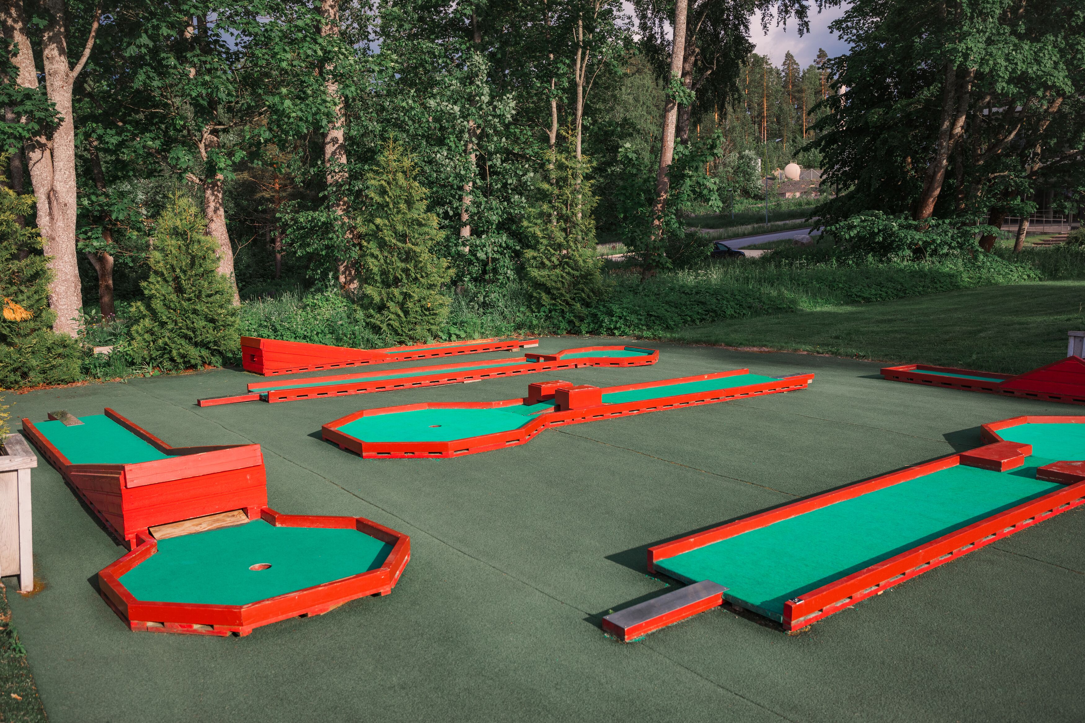 A miniature golf course with multiple red and green holes set in a grassy area surrounded by trees.