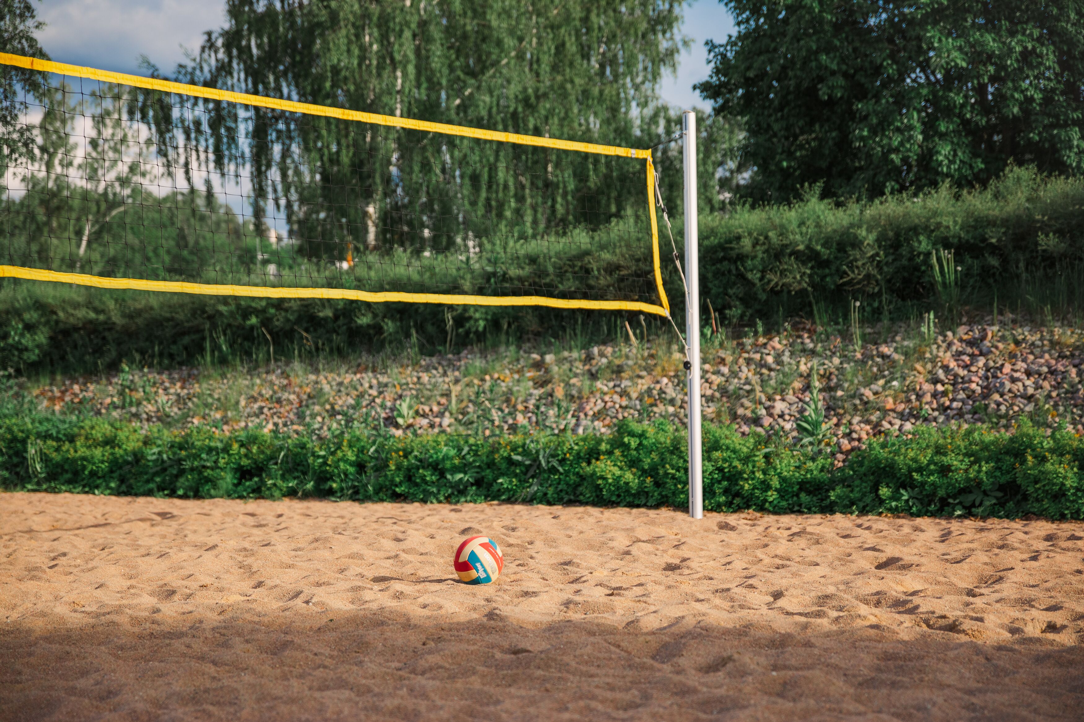 Un filet de volley-ball sur une surface sablonneuse, avec un ballon au premier plan, entouré de verdure et d’arbres.