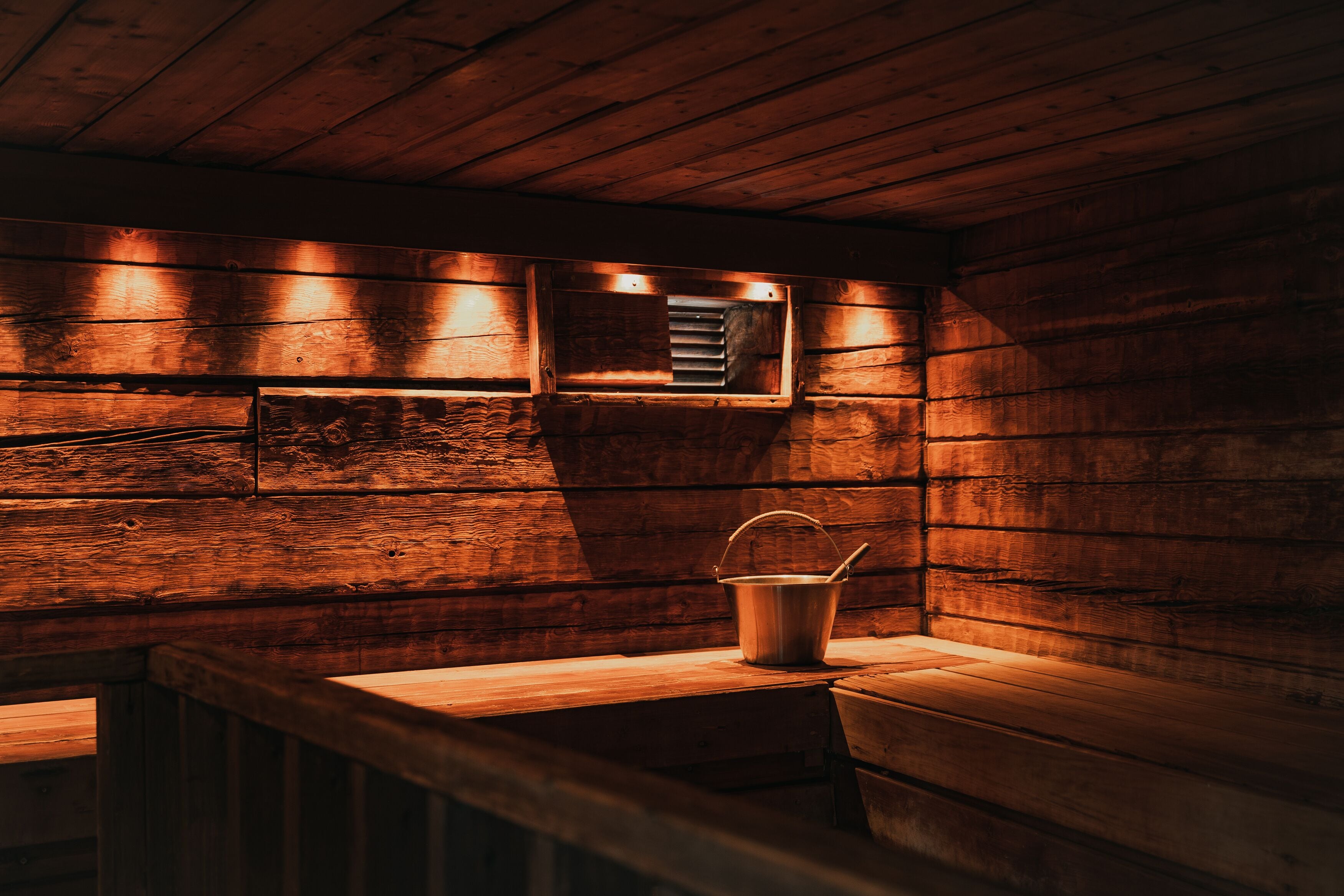 Interior de una cabaña de madera con un banco, un cubo metálico y un calentador fijado a la pared.