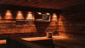 Interior de una cabaña de madera con un banco, un cubo metálico y un calentador fijado a la pared.