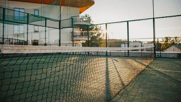 Een tennisbaan met een net, omringd door een gebouw met een gebogen dak en glazen hekwerk.
