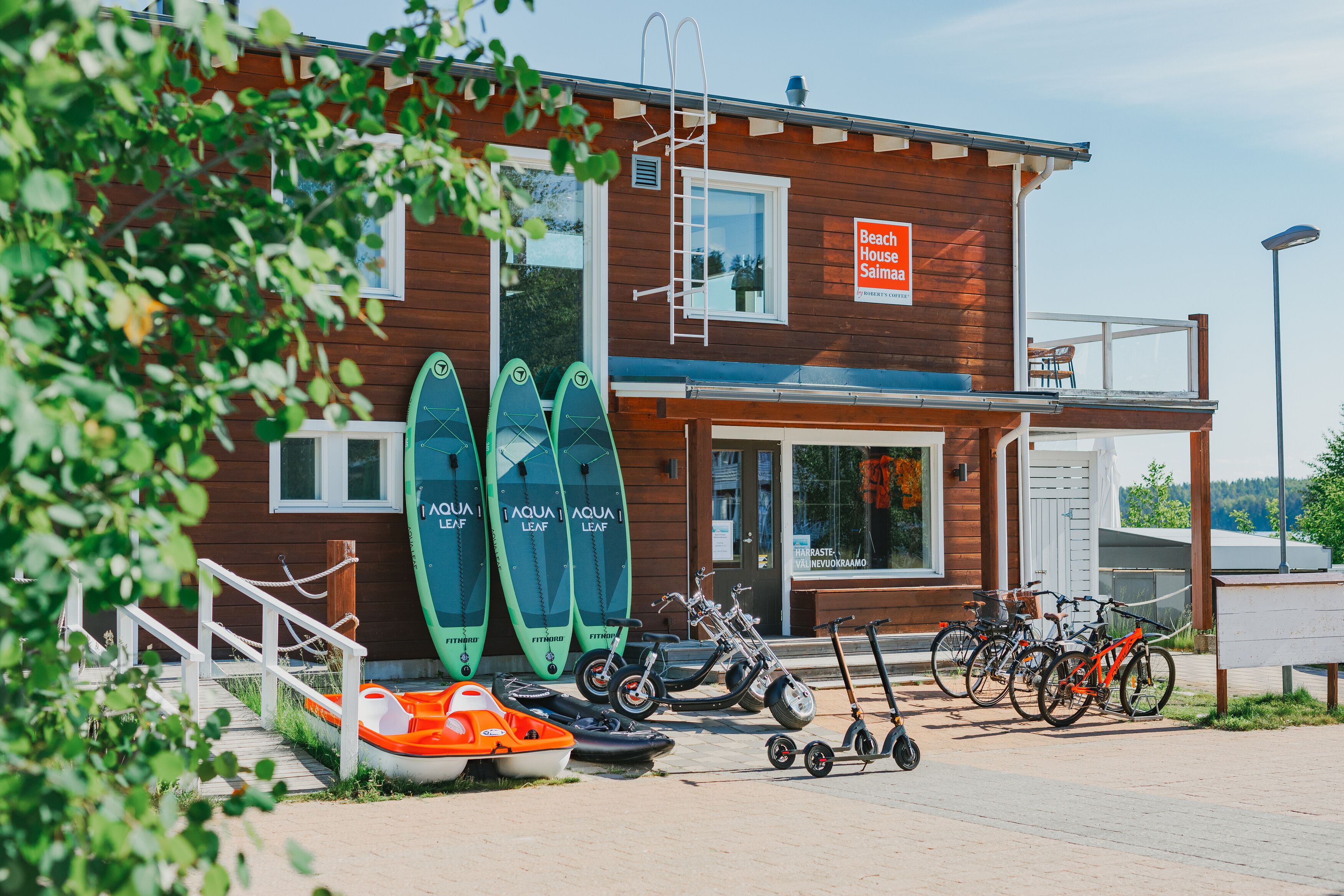 Il s’agit d’un bâtiment en bois sur lequel est apposée une enseigne indiquant « Beach House Salina ». On peut voir deux planches de paddle vert, un vélo et un scooter garés à l’extérieur.