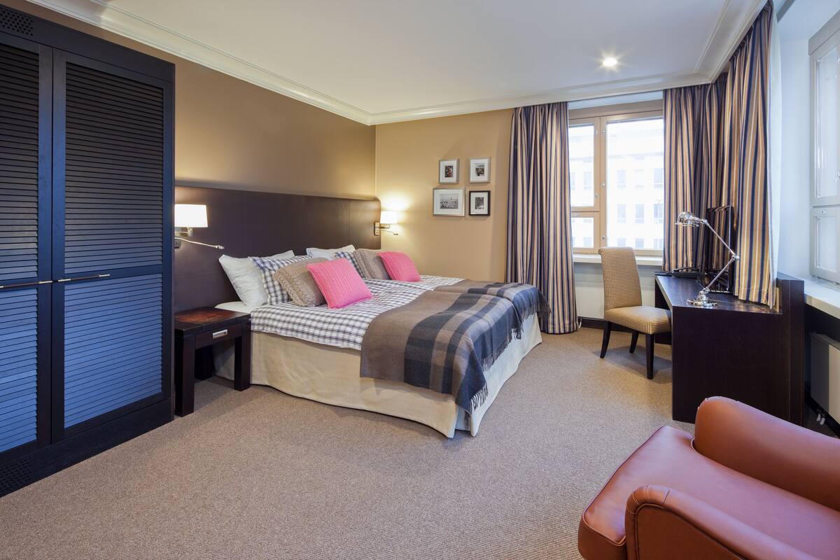 Une chambre d’hôtel comprenant un lit, une table de chevet, une lampe, une chaise, un bureau et une fenêtre avec des rideaux.