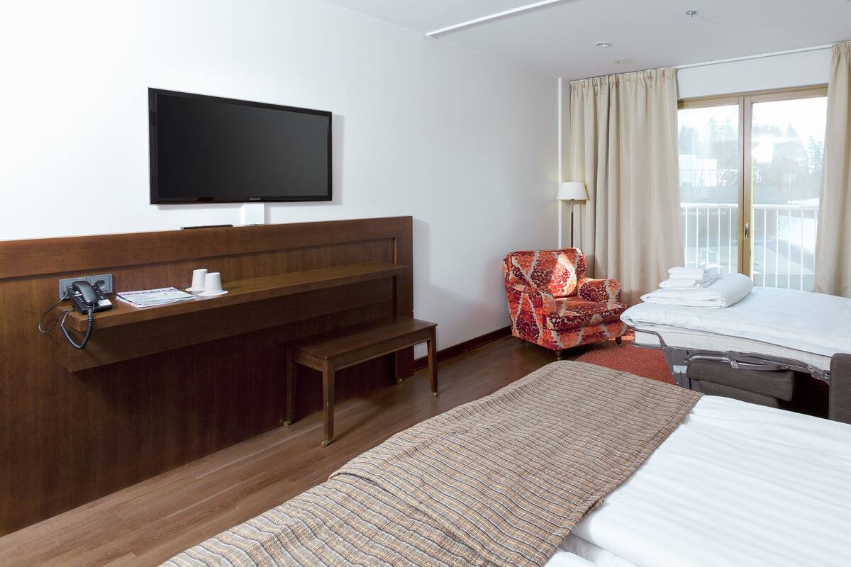 Une chambre d’hôtel avec un lit, un bureau avec un téléphone, une chaise, une télévision et une fenêtre avec des rideaux.