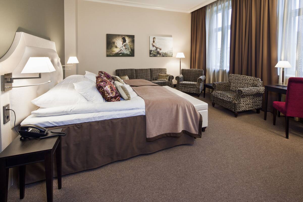 Une chambre d’hôtel avec un grand lit, deux fauteuils, un bureau avec un téléphone et une lampe.