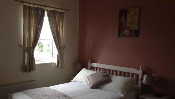 A bedroom with a bed, a nightstand, a window with curtains, and a wall-mounted painting. - Deerpark Manor Bed & Breakfast (Swinford)
