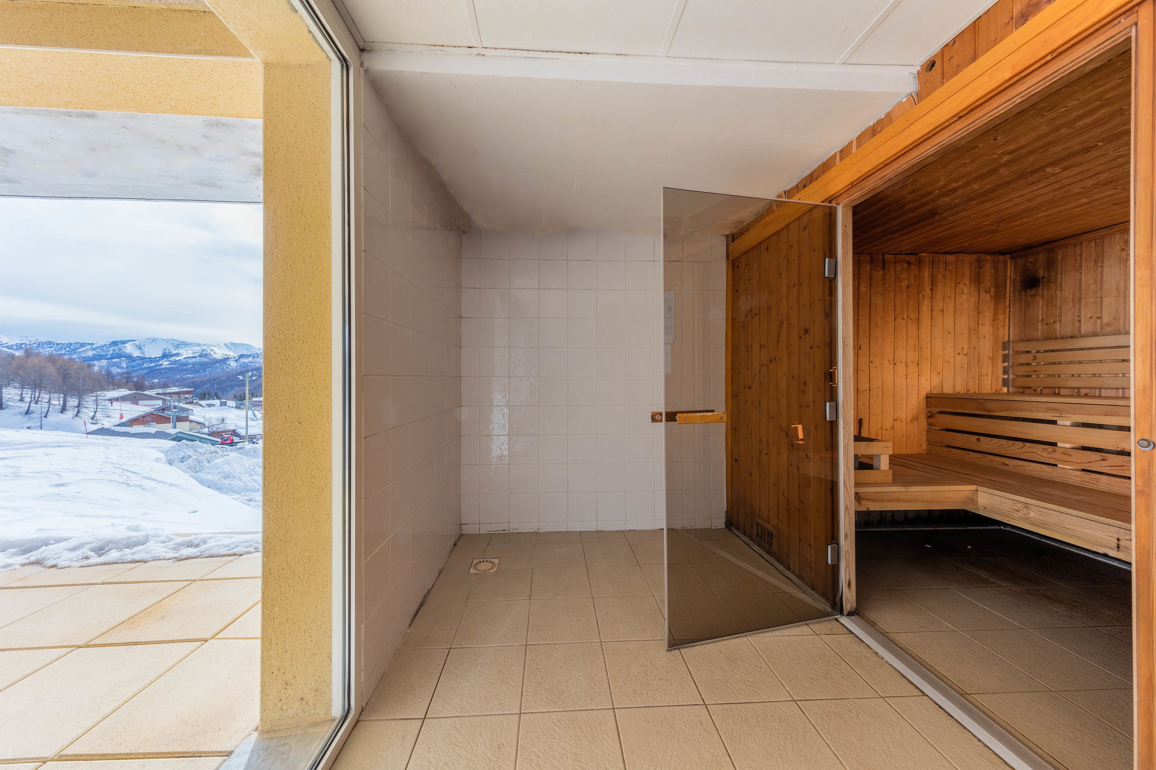 A sauna room with a wooden bench, a glass door leading to a snowy landscape, and tiled walls.