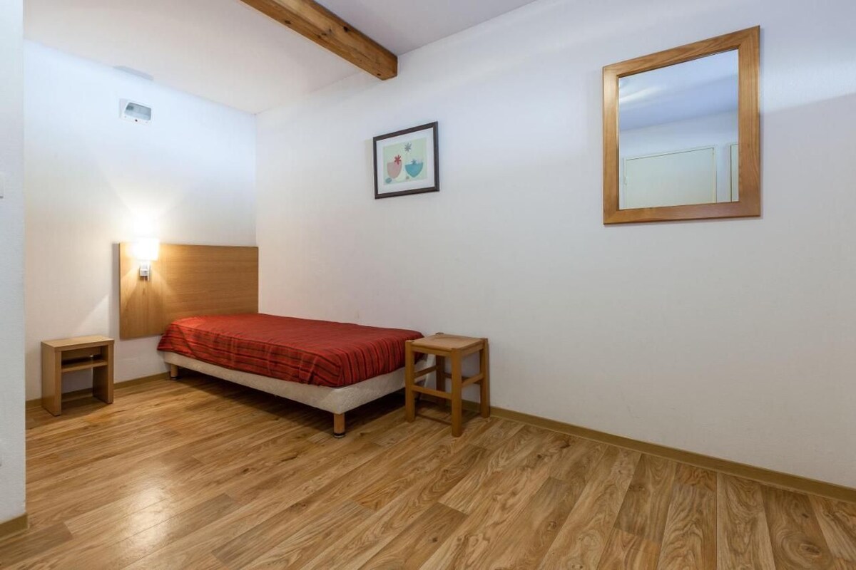 A bedroom with a bed, wooden floor, a headboard, a bedside table, a nightstand, a wall-mounted mirror, and a framed picture.