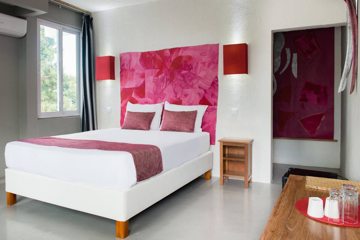 A modern hotel room with a large bed, a small wooden nightstand, a red and white wall art piece, and a window with curtains.