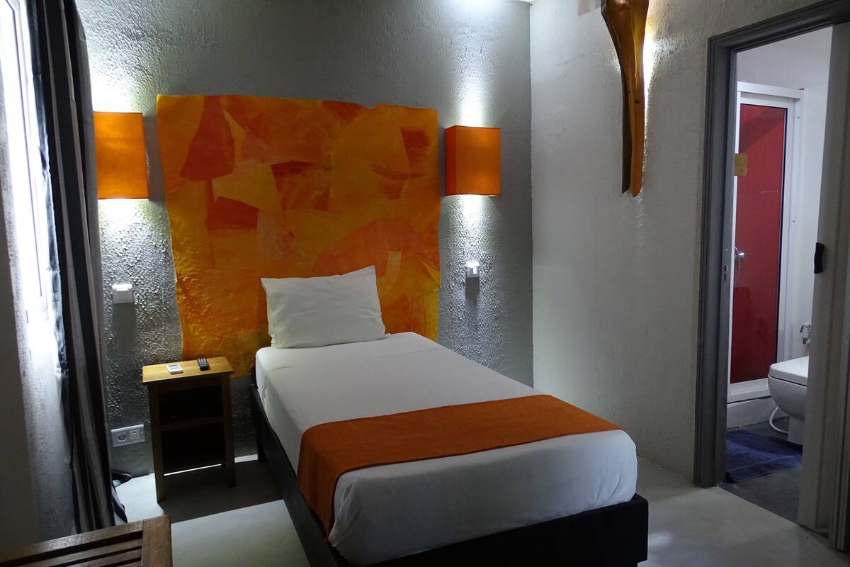 A hotel room with a bed, a nightstand, a wall-mounted lamp, and a door leading to a bathroom.