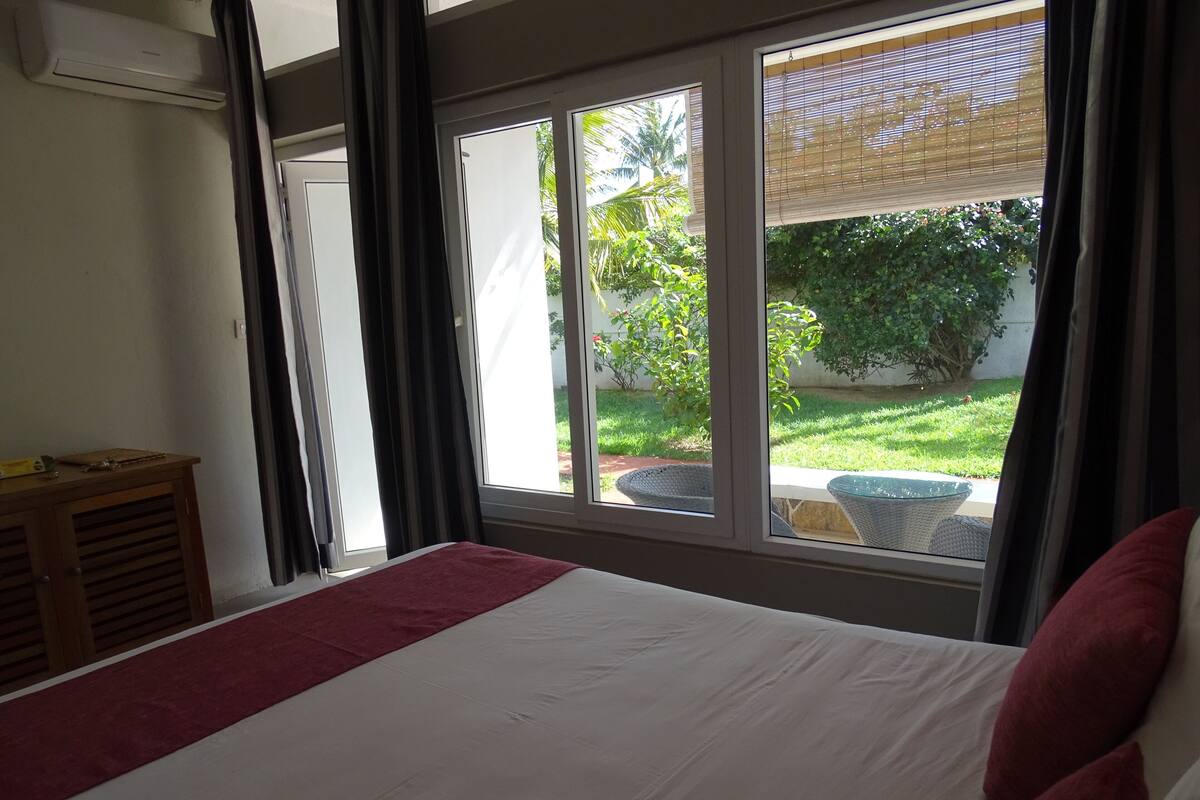 A bedroom with a large bed, a wooden nightstand, and a view of a garden through a window.