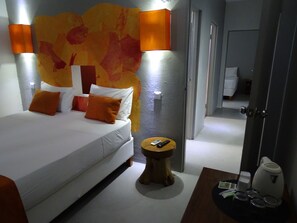 A modern hotel room with a large bed, two orange pillows, a bedside table with a remote control, and a small round table with a kettle and cups. - La Tonnelle (Trou aux Biches)