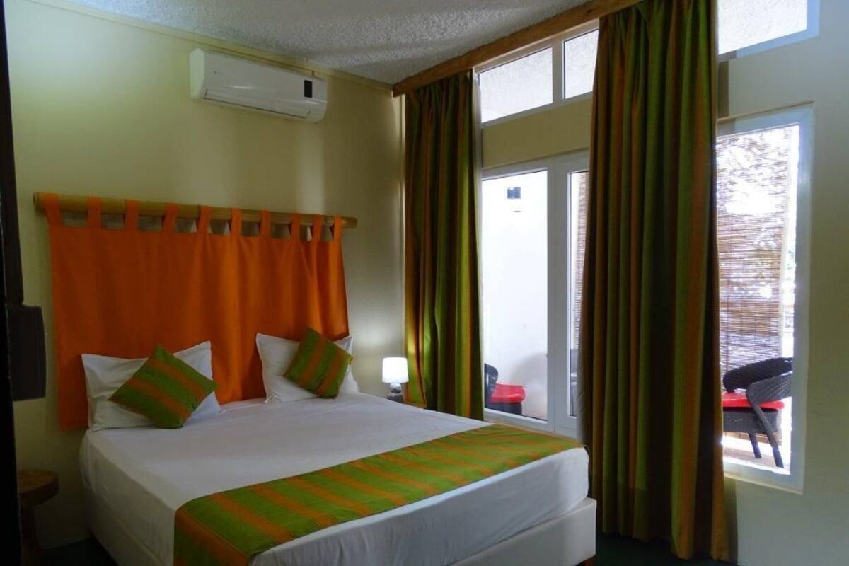 A hotel room with a large bed, a window with curtains, a door, and a small table with a lamp.
