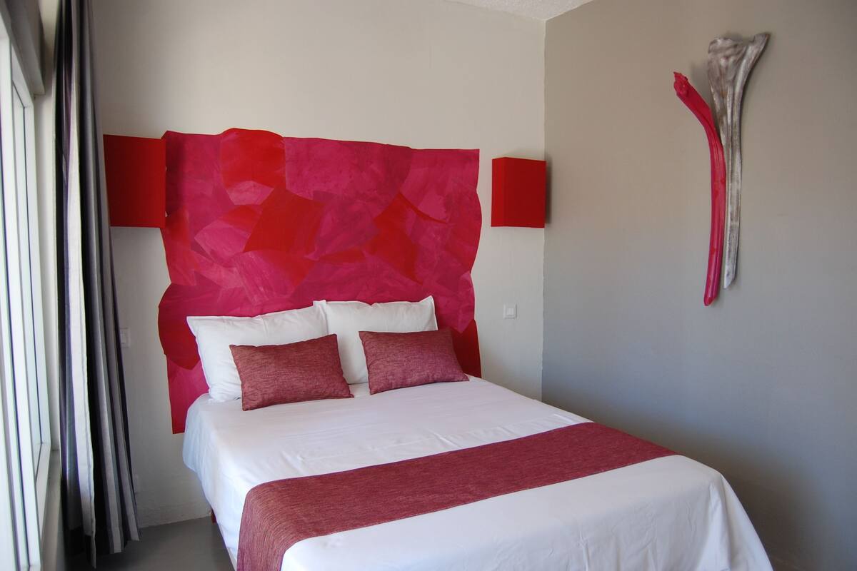 A hotel room with a bed, red and white bedding, a red headboard, and a wall-mounted sculpture.