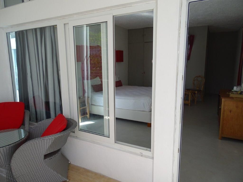 A room with a bed, a chair with red cushions, a wooden dresser, and a sliding glass door leading to another room.