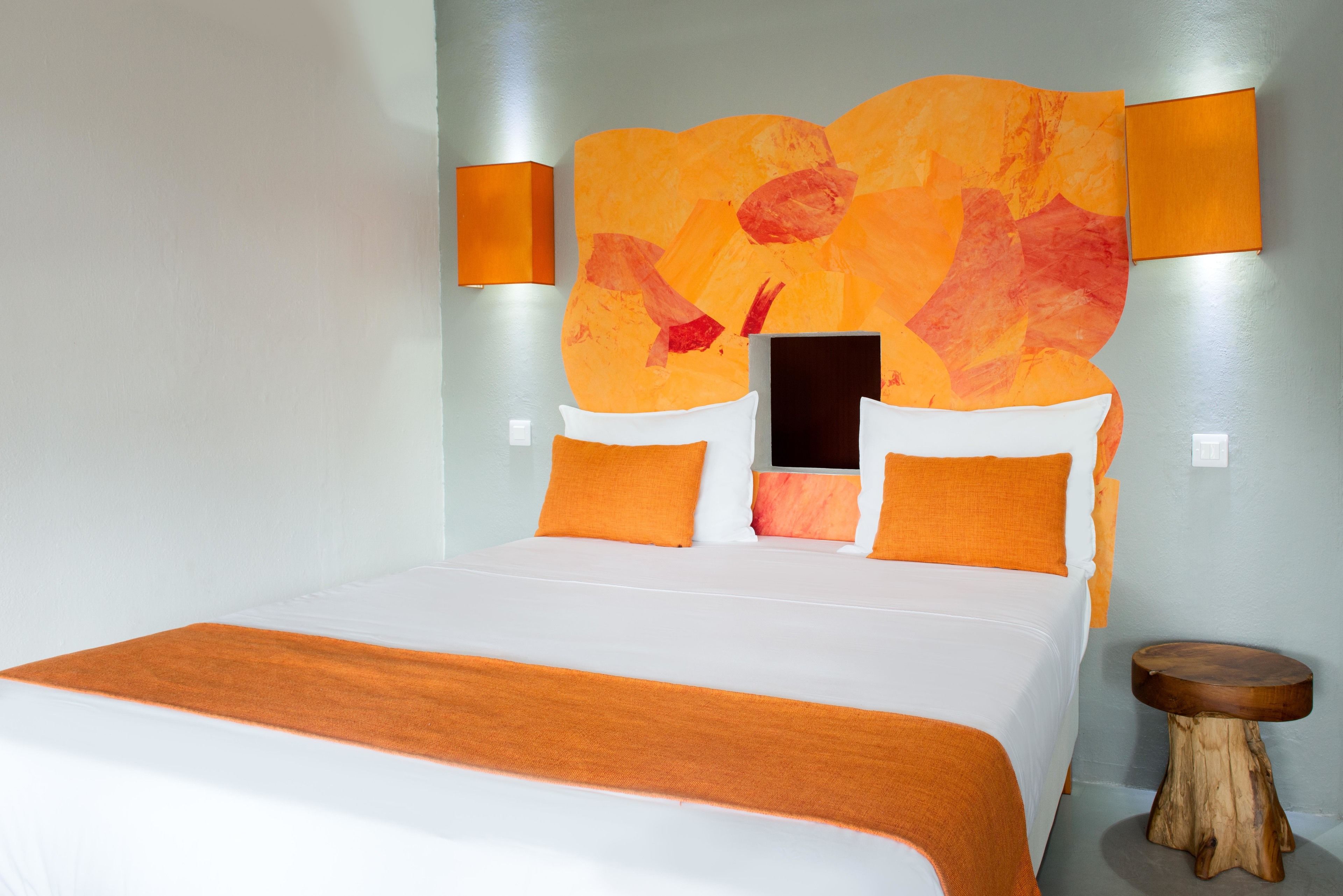 Una cama con colcha naranja, una mesita de noche de madera y una pared con un cuadro abstracto.