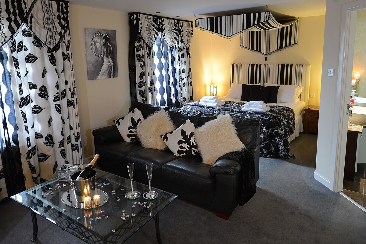 A modern hotel room with a black leather sofa, a glass coffee table with a candle, and a bed with striped curtains.