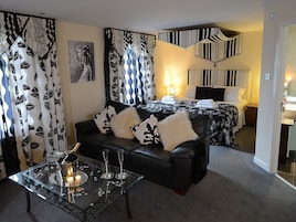 Una habitación de hotel moderna con un sofá de cuero negro, una mesa de centro de vidrio con una vela y una cama con cortinas a rayas.