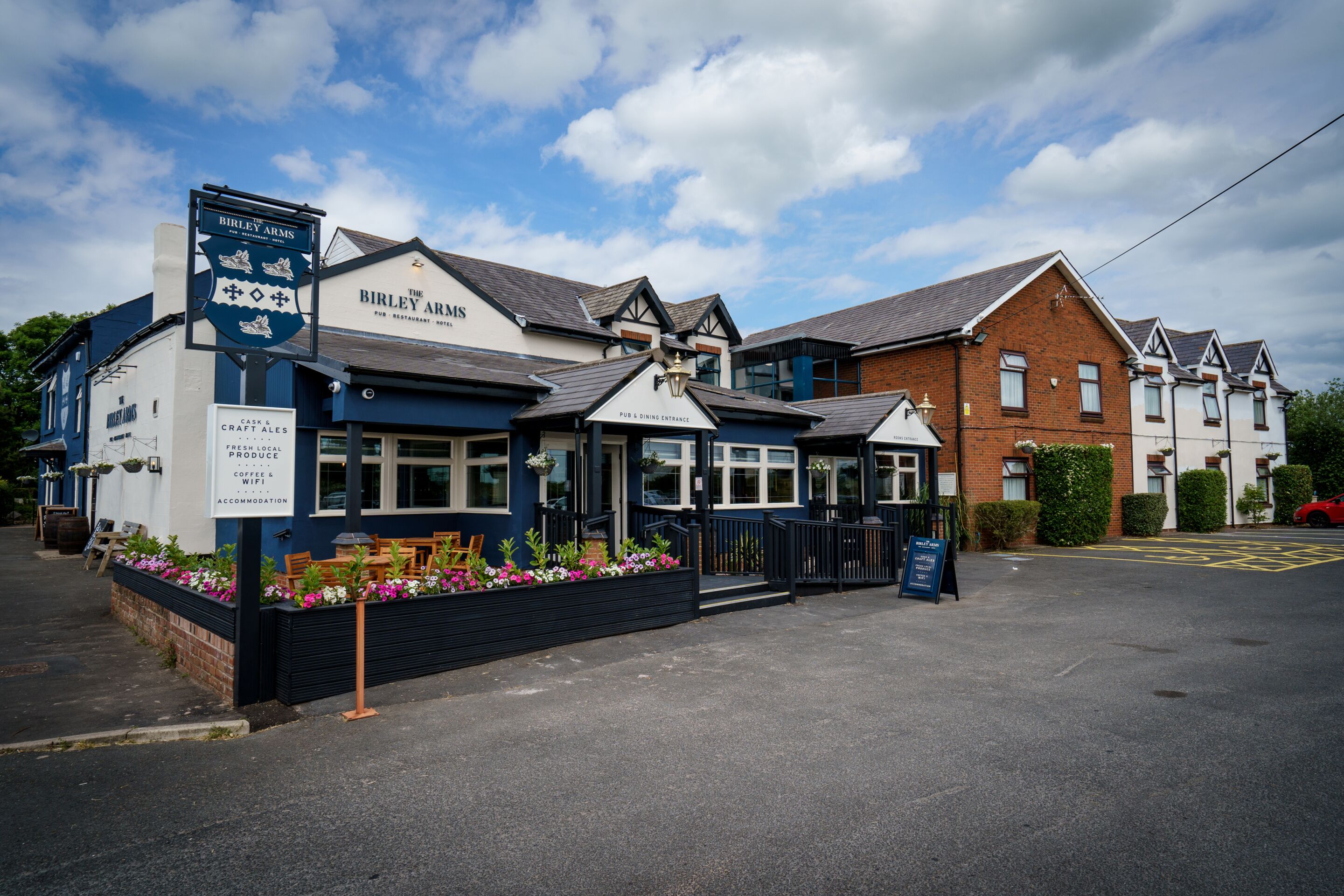 Pub chamado 'The Birley Arms' com área externa para sentar e placa anunciando cervejas em barril e artesanais, produtos frescos da região, café e wifi, além de hospedagem.