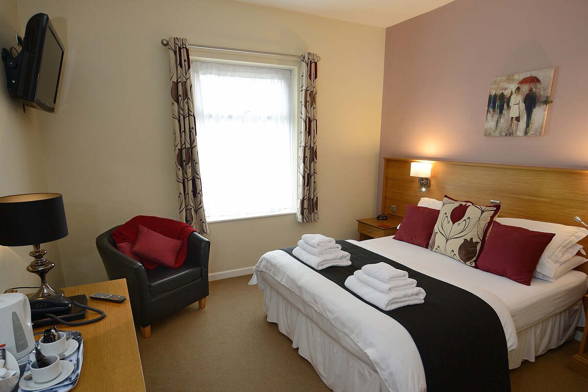 A hotel room with a bed, a desk with a lamp, a chair, a window with curtains, and a painting on the wall.
